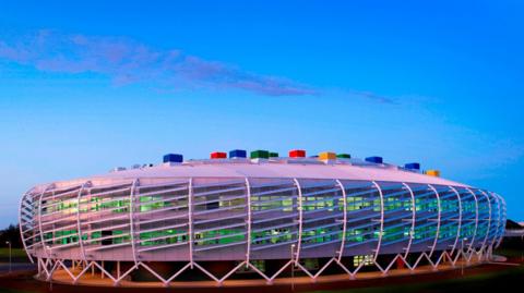 Colourful, oval-shaped, futuristic-looking Monkseaton High School which has multi-coloured blocks on its roof. The building is wrapped by white steelwork which sits on A-shaped white steel rods. Lights visible through windows make the rooms appear green coloured.