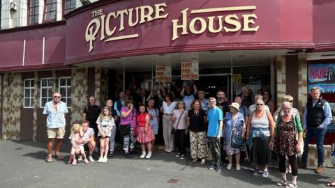 A large group of people of all ages standing outside a cinema building, some of them holding placards saying 'Save Our Cinema'. The big sign on the building identifies it as "The Picture House".