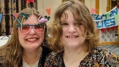 A woman and her teenage daughter smiling at the camera. The woman is wearing pink flamingo sunglasses, a dark coloured cardigan and white top. The teenager has shoulder-length curly hair and is wearing a black and orange pattered shirt. There is colourful bunting and banners in the background.