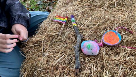 Brightly painted pebbles and twigs with coloured wool tied on have been placed on a hay bale. A child's hands are visible on the left. There is grass in the background.