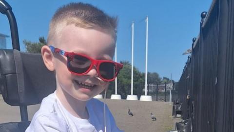 Dáibhin McConnell-Mailey, a four-year-old boy with short, blond hair, smiles at the camera while sitting in a wheelchair. He has red sunglasses on.