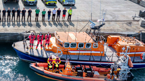 A group of emergency service personnel stand in a line near the edge of a dock. There are two boats pulled up to the dock, the one closer to the wall is bigger. Three people stand in orange jumpsuits on the deck of the larger vessel. Two people in yellow and black overalls stand on the deck of the smaller boat. They are all looking at the camera.