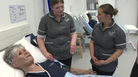 The image shows a clinical training ward where healthcare students in uniform are gathered around a hospital bed, practising care on a lifelike patient mannequin connected to monitoring equipment. The space is bright and modern, with another simulated patient visible in a bed behind them, reinforcing the realistic hospital setting.