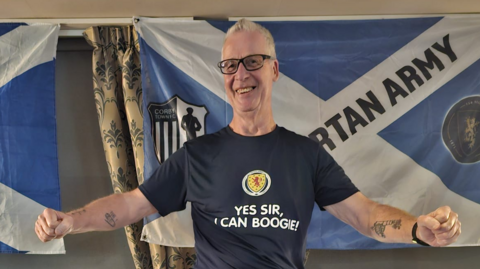 A man in glasses and a navy T-shirt stands in front of two Scotland flags, smiling. He is wearing glasses and a dark blue shirt which has the Scotland badge on it and the words "Yes sir, I can boogie!"