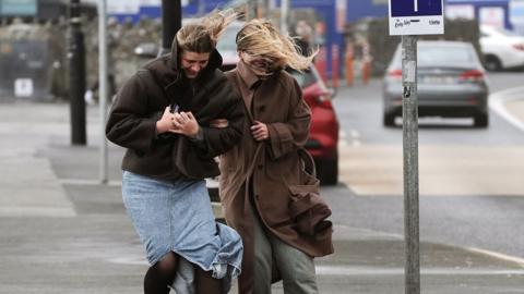 two women battle the wind whilst walking down a street.