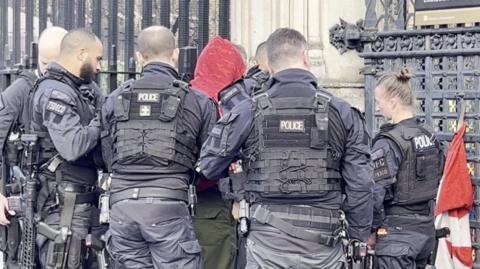 Police officers pictured wearing navy uniform and black body armour arrest a man wearing a red hood. They are standing in front of black gates at the entrance to the Palace of Westminster.