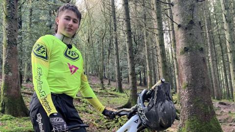 A man is stood in a forest next to a mountain bike. He is wearing a bright yellow jersey, black trousers and black and white gloves. He has brown hair in a mullet style. He is looking at the camera with a slight smile. 