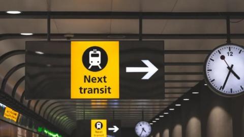 A yellow sign with the words "Next transit" written in black. The sign is hanging from the ceiling inside a tunnel. To the right is a white clock face showing the time 16:34.