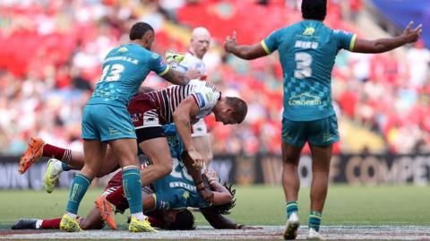 Wigan Warriors player Sam Walters, in cherry and white hoops, dumps Hull Kingston Rovers player Bill Leyland on the ground head-first, in a tackle made with team-mate Junior Nsemba, partly hidden. Hull KR players Eliot Minchella and Peta Hiku look on.