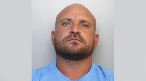 A police mugshot of Ashley Hunt. He is bald with a dark brown beard and a neutral facial expression. He is wearing a light blue t-shirt and standing against a grey background.