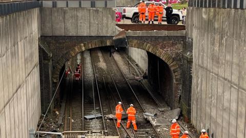 Workers are seen clearing up the damage on the Manchester-Liverpool rail line; above there is a bridge with a part missing where the car veered off the road above.