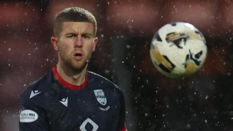 Ryan Leak in action for Ross County. He is wearing a dark blue shirt and is watching the ball move away from him after sending a long pass forward.