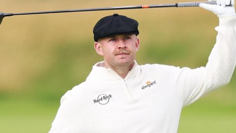 Harry Hall, wearing a flat black cap, with a golf club in his hand