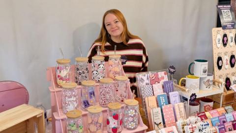 Alaina Davies smiles at the camera as she stands behind a stall that is displaying her products, such as colourful water bottles, mugs and keychains. She has auburn hair and is wearing a striped jumper. 