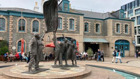 Liberation Square in St Helier, Jersey. A statue at the centre of a fountain shows a group of people celebrating the island's liberation from Nazi forces during World War Two. A building called Liberty Wharf is behind the statue. Several people are walking around the square or sat on tables outside a restaurant.