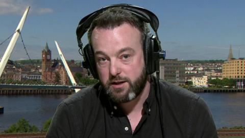 Colum Eastwood sitting in a studio. There's a backdrop of Foyle behind him. He's wearing head-phones