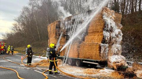 Fire fighters are seen extinguishing a lorry fire with hoses. The lorry is on the M5 with hay bales four stacks high.