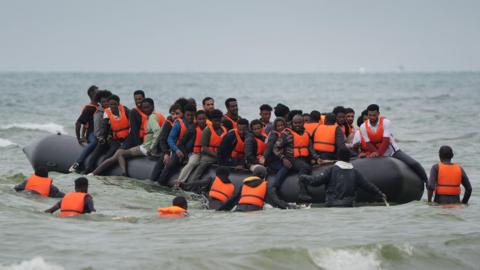 A crowded inflatable boat carrying migrants in choppy coastal waters, with several people in life jackets entering or surrounding the vessel.