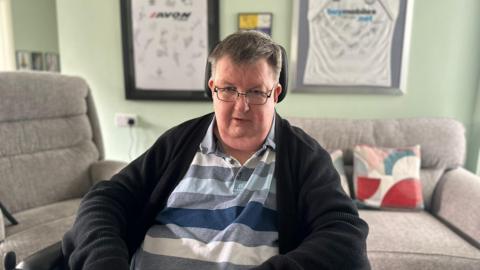 A man with grey hair and glasses whilst being sat in a wheelchair inside his living room