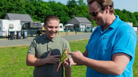 A boy smiling and looking down towards a parrot-type bird being held by a man in a blue polo-shirt wearing dark glasses and smiling and looking at the bird