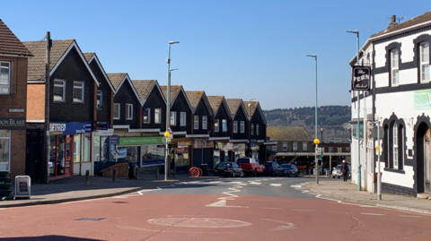 The view of a high street. There's a roundabout and a zebra crossing on the road. There's a row of shops on the street. Cars are parked further along the road. The sky is clear and blue, hills can be seen in the background. 