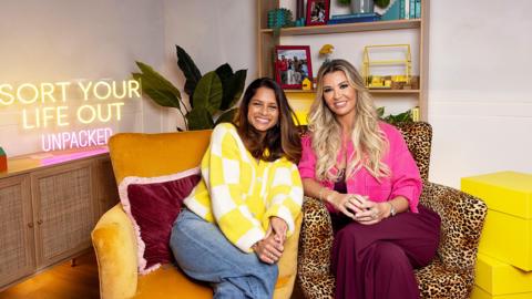 Dilly Carter and Christine McGuinness smiling indoors in front of a Sort Your Life Out Unpacked sign
