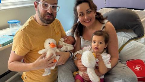 A man and a smiling woman are seated on a hospital bed. The man is holding a newborn and a duck toy, the woman has on her lap a toddler.