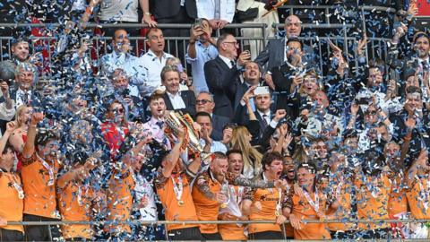 John Egan of Hull City in an orange playing shirt lifts the trophy after Hull City win the Championship play-off final with the players being showered in ticker-tape