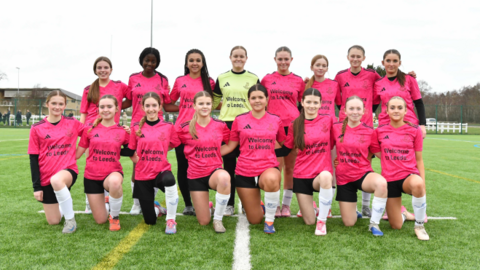 A group picture of girls on a football pitch wearing matching kits, which consist of pink t-shirts, black shorts and white socks. They are split into two rows with several stood up and the rest kneeling down in front.