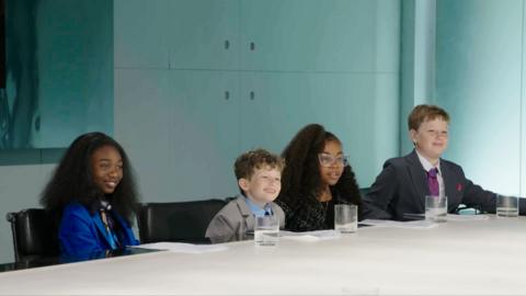 A group of four children in formal attire sit at a conference table, with glasses of water, in a modern meeting room.