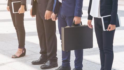 A row of four people, seen from the waist down, two of whom are in leggings while the others are wearing brown or blue trousers. One is carrying a black briefcase and two others are carrying iPads.  They are standing on square white slabs.