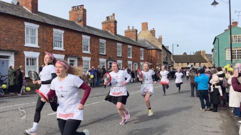 A group of women in aprons and themed outfits run down a street while holding frying pans with pancakes, taking part in The Olney Pancake Race. Spectators line the roadside, watching and taking photos in a residential neighborhood.
