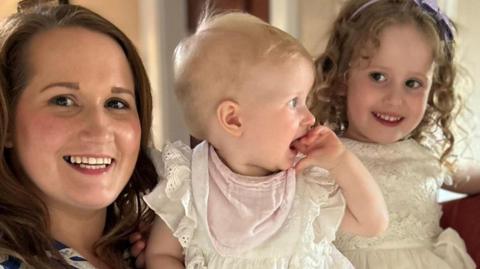 A woman with dark brown hair is looking at the camera and smiling. She is holding a blond baby in her arms, the child is wearing a cream top, pink bib and has her hand in her mouth. Beside her is another young child with shoulder-length dark blonde curls. She is wearing a cream dress and looking off to the side of the image. 