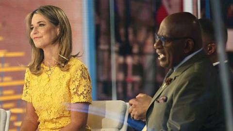 Savannah Guthrie, in a bright yellow dress with a lace floral pattern, smiles as she sits next to Al Roker on the "TODAY" show set