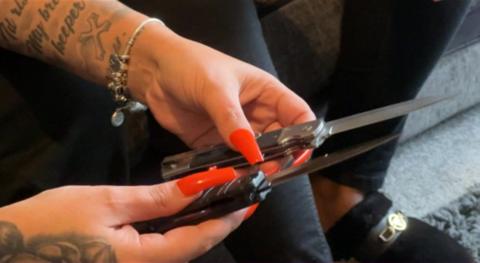 A woman with long red nails and tattoos on her arms holding two pocket knives