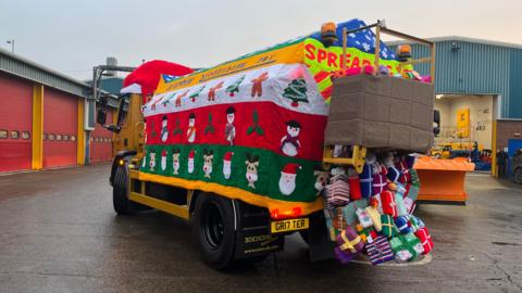 A yellow gritter covered in a "knitted jumper". Pictures of gingerbread men, reindeer and Santa make up the design, as well as knitted presents spilling out of a sack at the back.