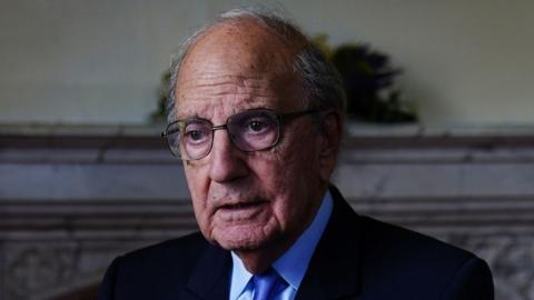 George Mitchell. He has grey hair at the sides, wearing glasses, a navy blazer, light blue shirt and blue tie. He is holding his hand out to the front. A marble fireplace is behind him.