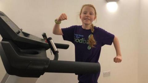 Luna raises a fist at the camera as she walks on an inclined treadmill in her house. She has red hair in a ponytail and wears a purple t shirt with Bristol North West Foodbank written on it