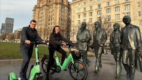 A man is on a green Bolt e-scooter and a woman is on a green e-bike. They are posing for the camera at Liverpool's waterfront in front of the Royal Liver Building and The Beatles statue