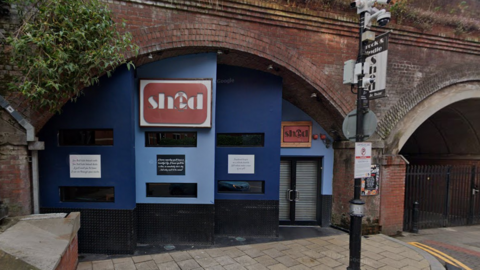 A blue building underneath railway arches with a brown sign reading: Shed