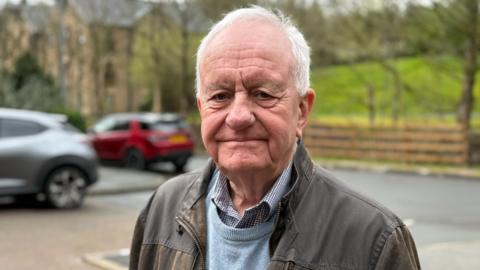 Steve Hindley is pictured outdoors near a residential street. He has short white hair and a lined face and is wearing a brown jacket over a light blue jumper and check shirt. His face is turned toward the camera with a slightly sad expression and a closed mouth. There are parked cars and a grassy hillside softly blurred in the background, as well as a large three-storey house in brown stone and with a pitched roof.
