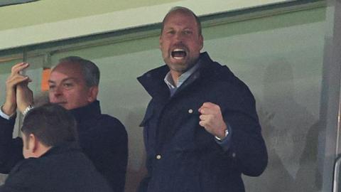 Prince William, wearing a blue coat, is cheering with his fist in the air at chest height
