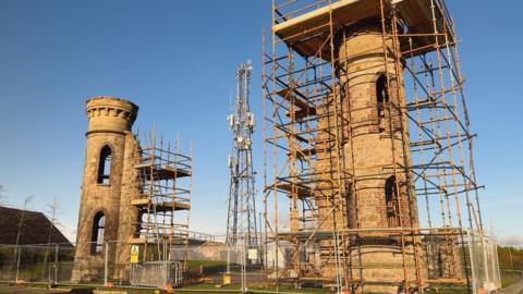 Two large stone pillars stand a distance apart in a green area. They are both surrounded by scaffolding and metal fences. In the middle is a large metal pylon.