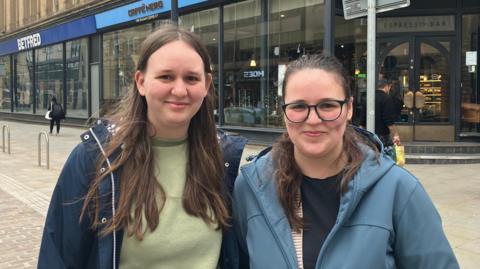 Two women with long brown hair pose together on a city centre street