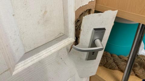 A white wooden door which has been damaged. The silver handle of the door hangs off.