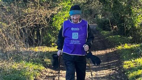 Derrick Downs walking with sticks towards camera through a woody glaze in bright sunshine wearing a woolly hat and mauve bib