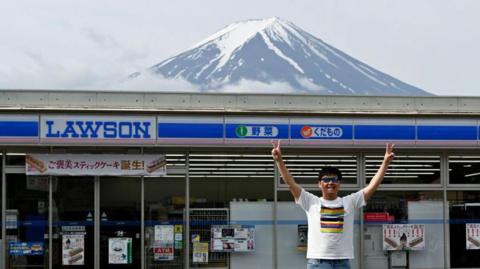 A tourist poses for photograph with Mount Fuji in front of a Lawson convenience store