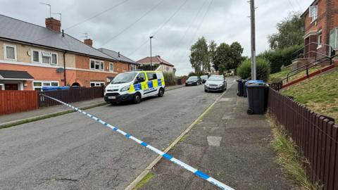 A police van is parked in a residential street with police cordon tape blocking the road. Houses and bins are pictured on either side of the road
