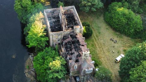 A drone image of Armathwaite Castle showing the damage caused by a fire. The building has a main rectangular structure with no roof and another structure connected to it, which is more similar to a house in shape. The roof is partially still on this section. Inside the affected areas are completely black with debris and collapsed beams and supporting structures. There is a river on one side. On the other side there is a lawn with an emergency vehicle parked on it and a group of people standing nearby.