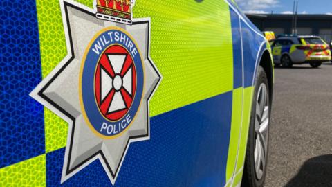 The side of a Wiltshire police car. It is parked in a car park.